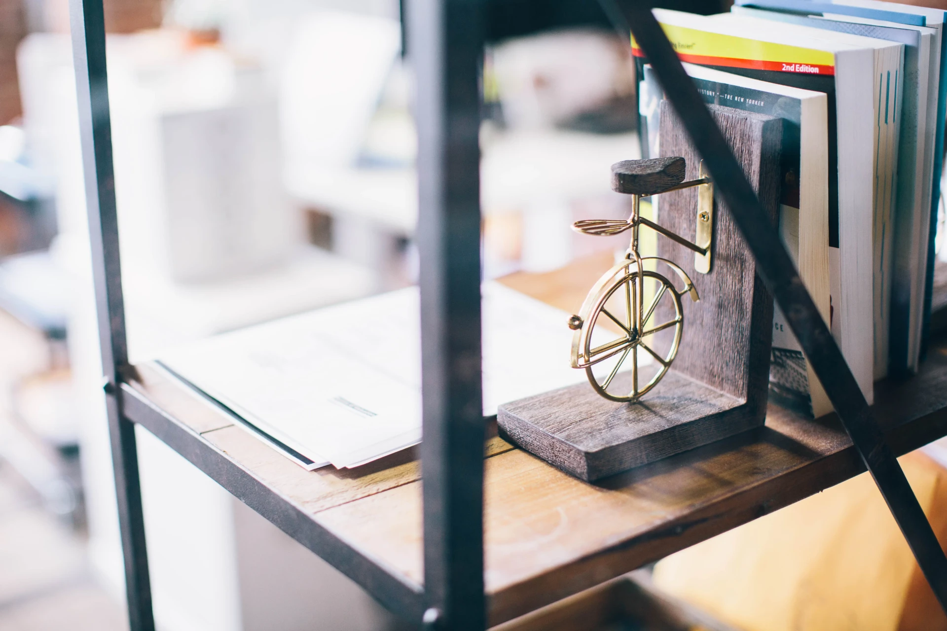 A wooden bookend with a small brass bicycle on an office shelf, holding a row of books beside a stack of papers